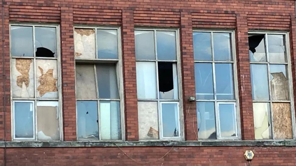 Red-brick wall of the East Mill with tall windows, their glass smashed and the panes patched with boards and peeling paper.